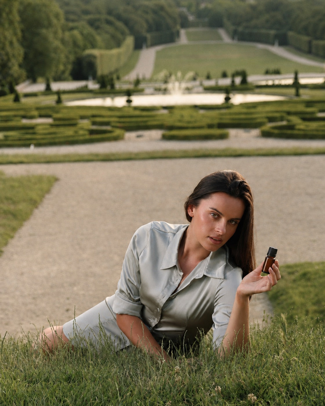 A woman resting on the lawn of a formal French garden, holding an amber bottle of Lavande Fine, with clipped parterre hedges and a fountain behind her.