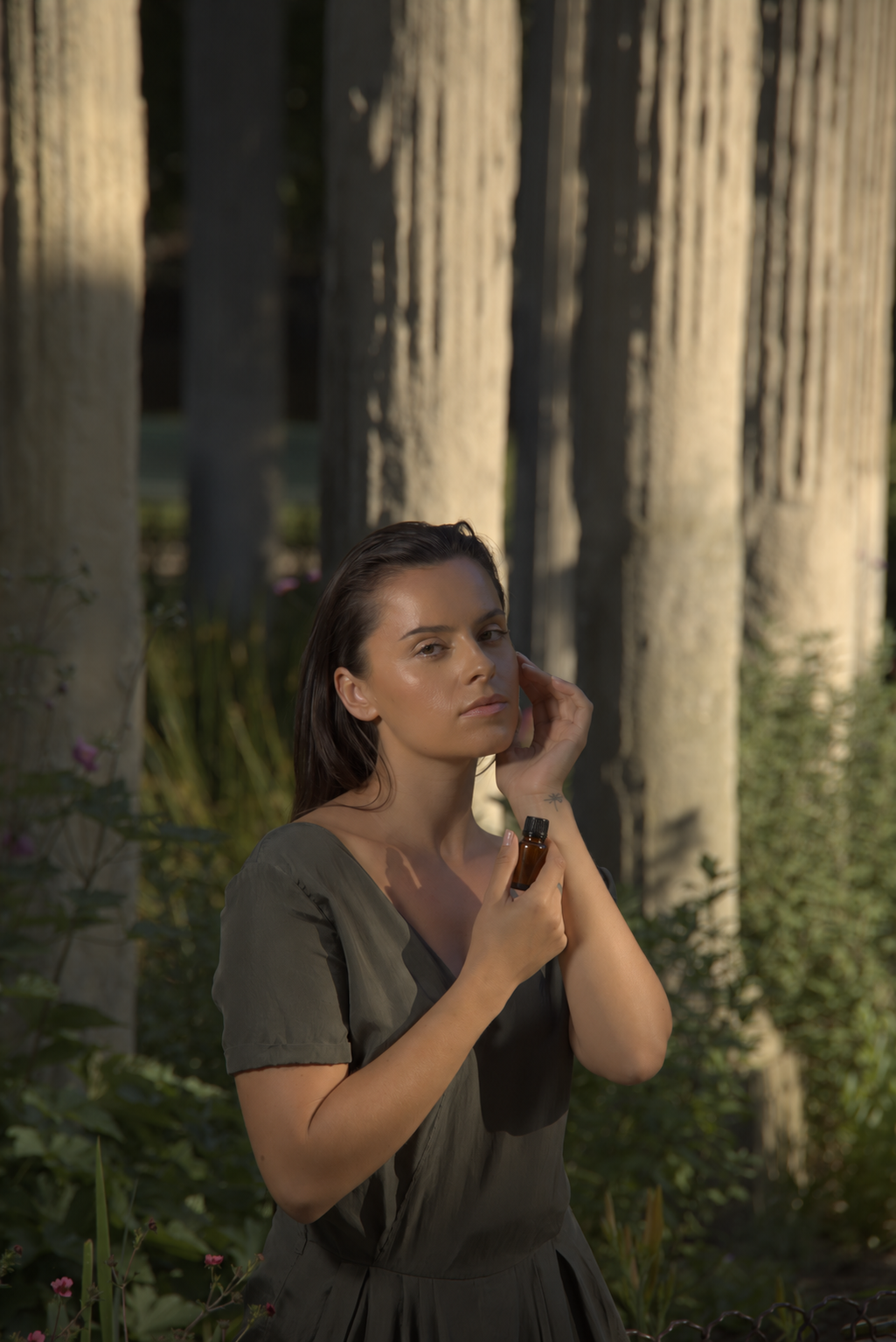 A woman in dappled sunlight beneath a colonnade, holding an amber bottle of rosemary oil to her neck.