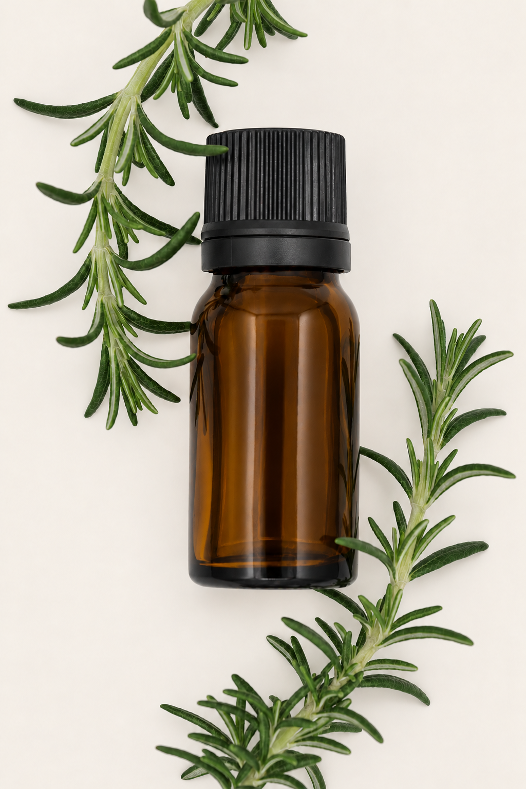 A flat lay of an amber glass bottle of rosemary essential oil resting among fresh rosemary sprigs on a cream background.
